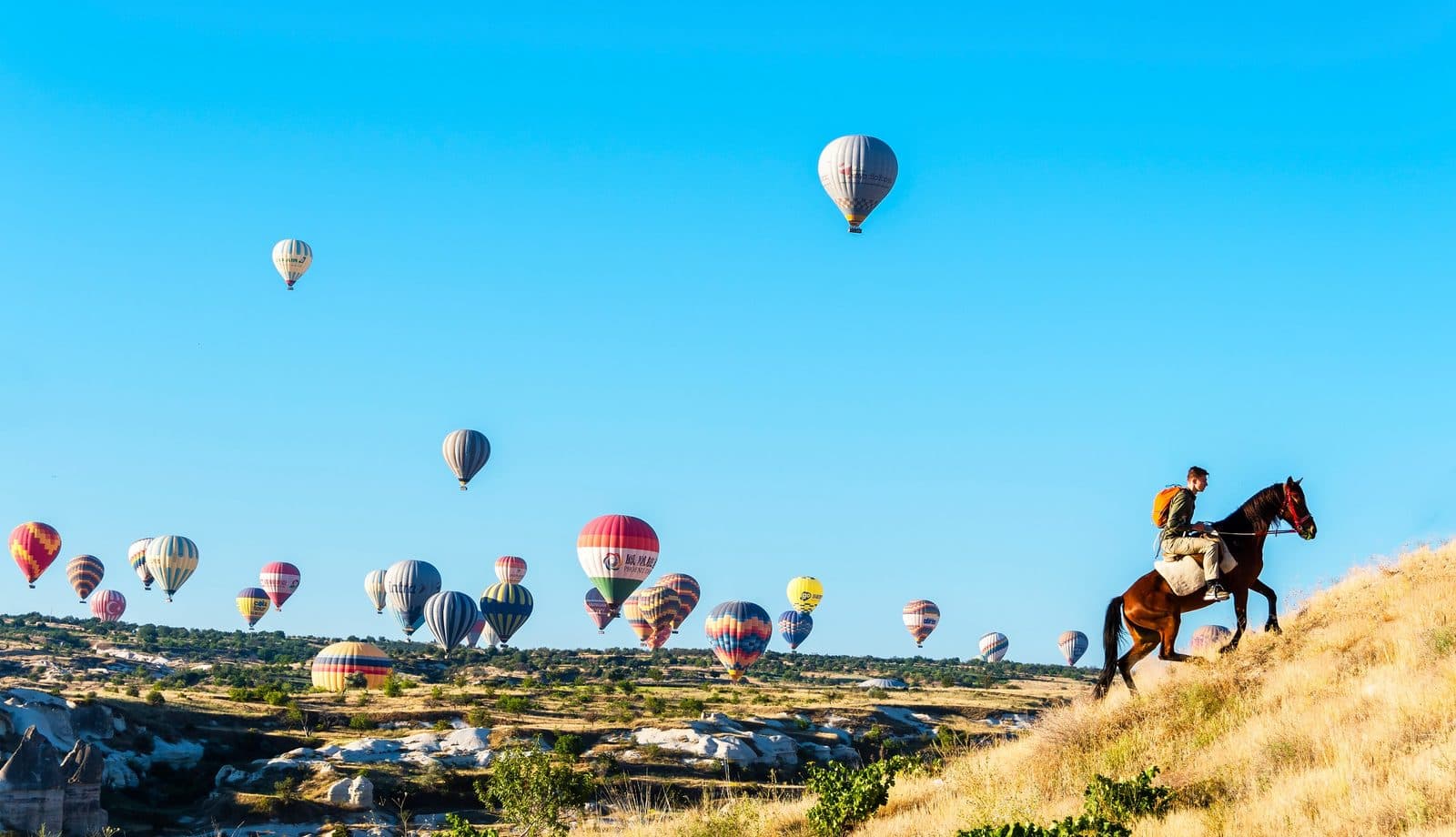 A rider on a ridge with hot air balloons filling the sky. Sunrise Horse Ride, arranged from The Owl Cave Hotel in Göreme, Cappadocia.