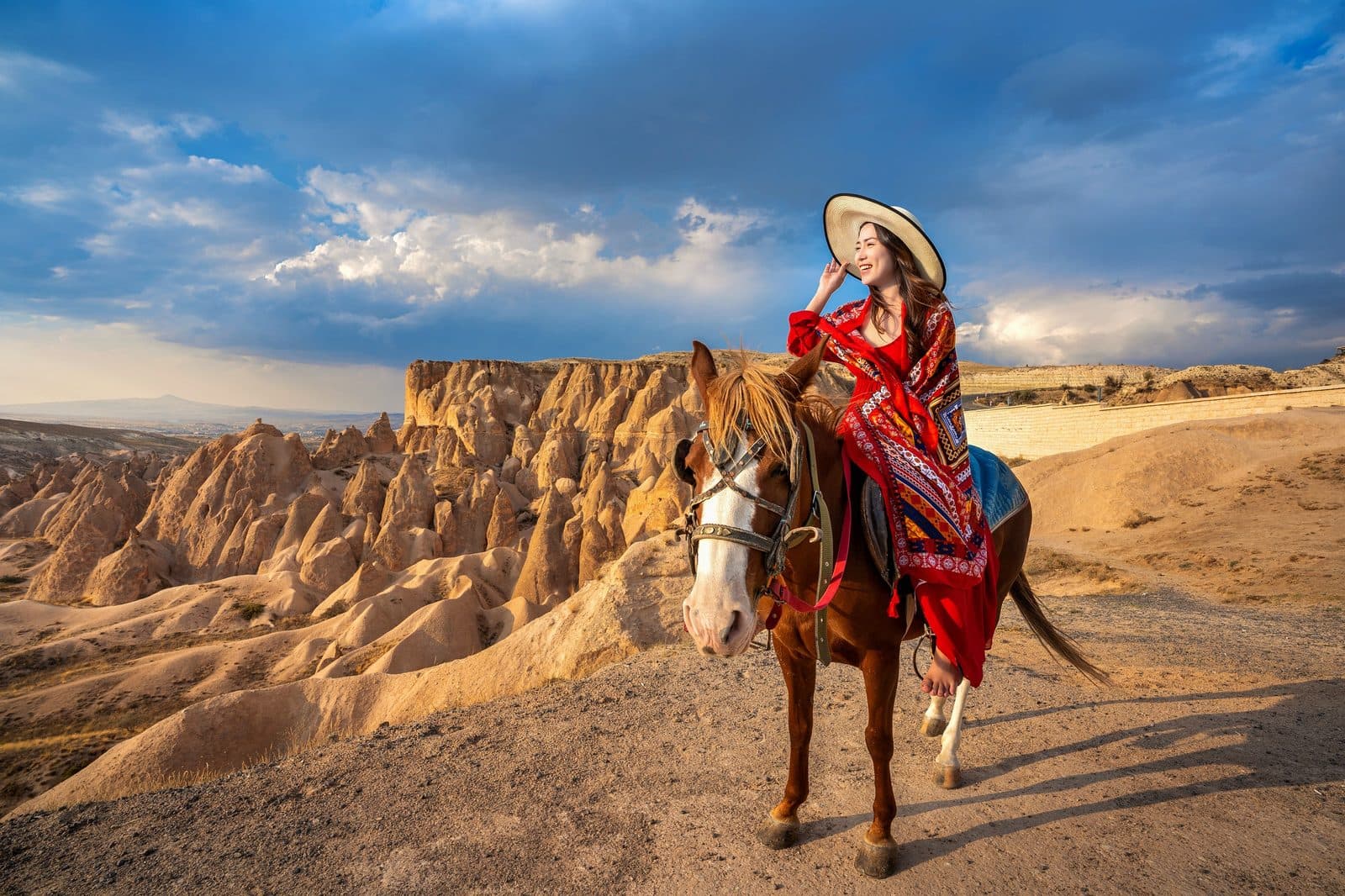 A rider in a red dress and hat on a brown horse, pink hills behind. Sunset Horse Trek, arranged from The Owl Cave Hotel in Göreme, Cappadocia.