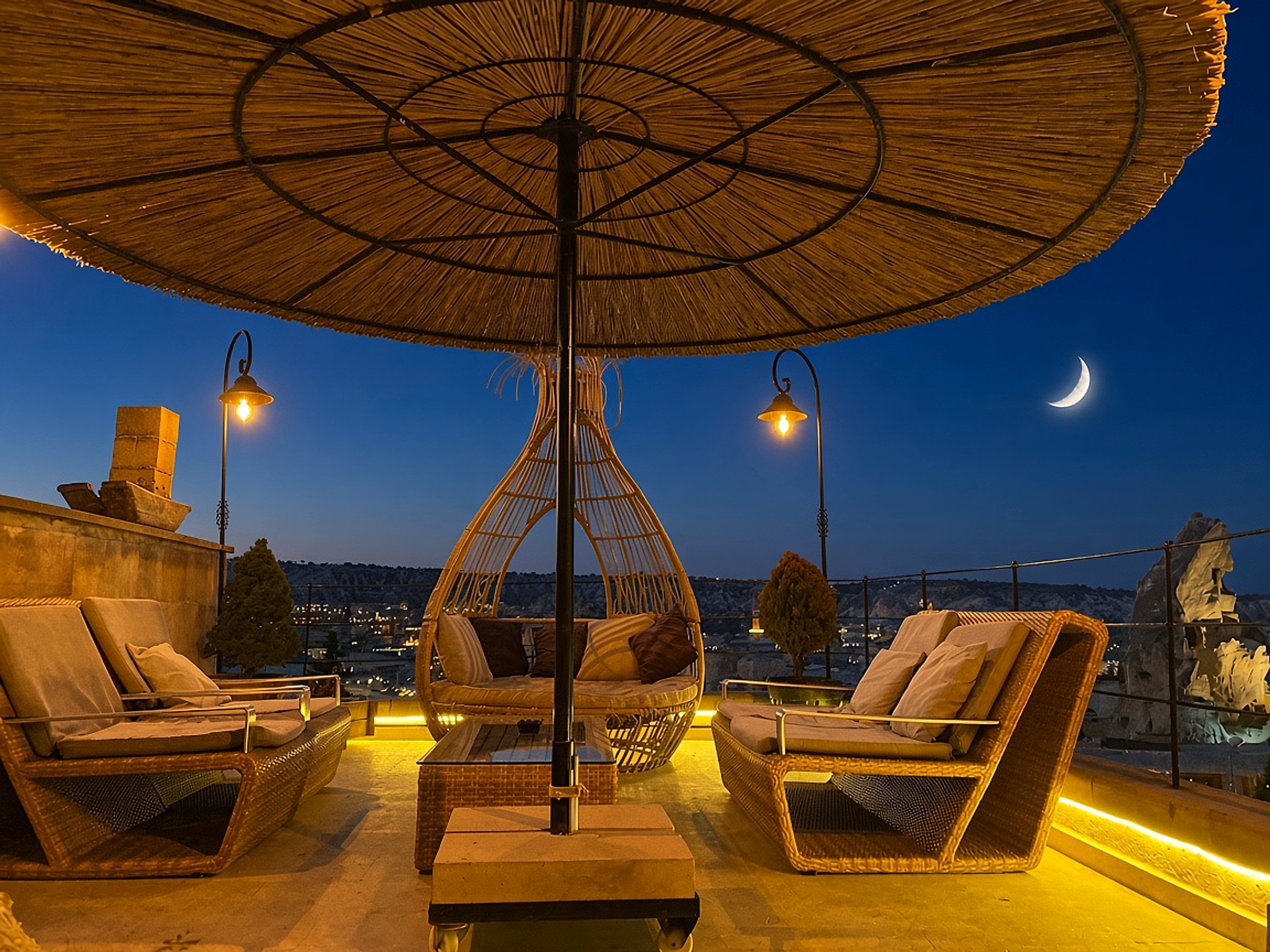 Dusk terrace at The Owl Cave Hotel in Göreme, with lounge chairs and a hanging egg seat under a thatched umbrella
