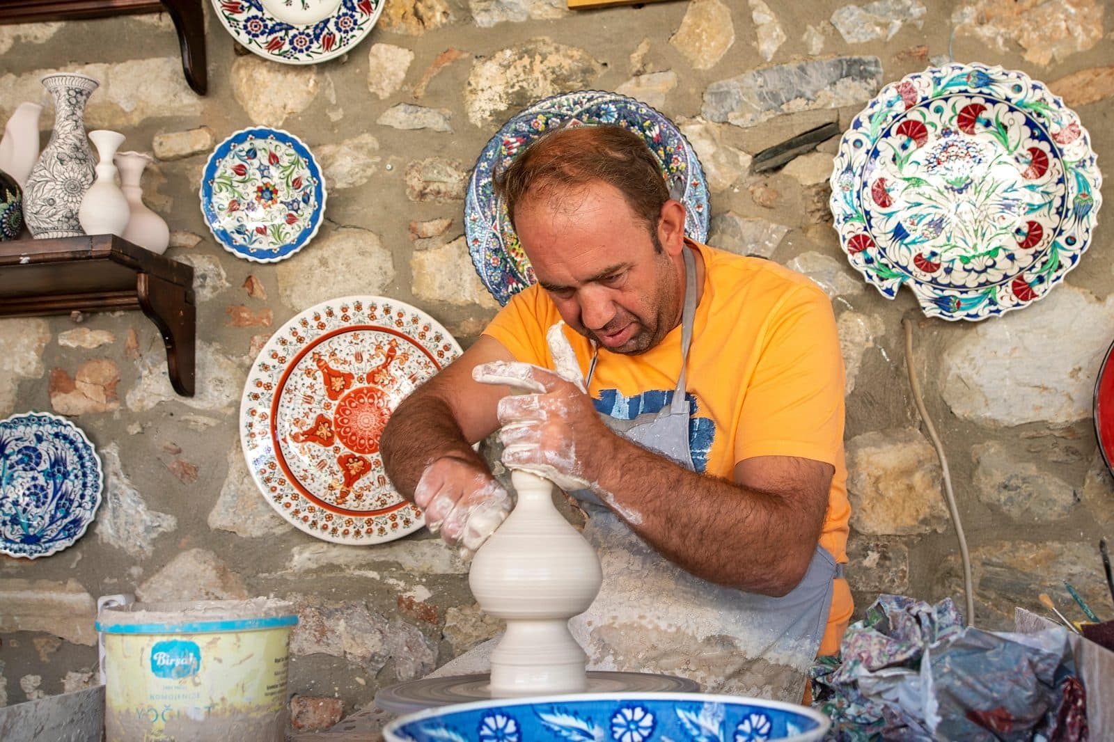 A potter throwing a pot, painted plates behind him. Avanos Pottery Workshop, arranged from The Owl Cave Hotel in Göreme, Cappadocia.