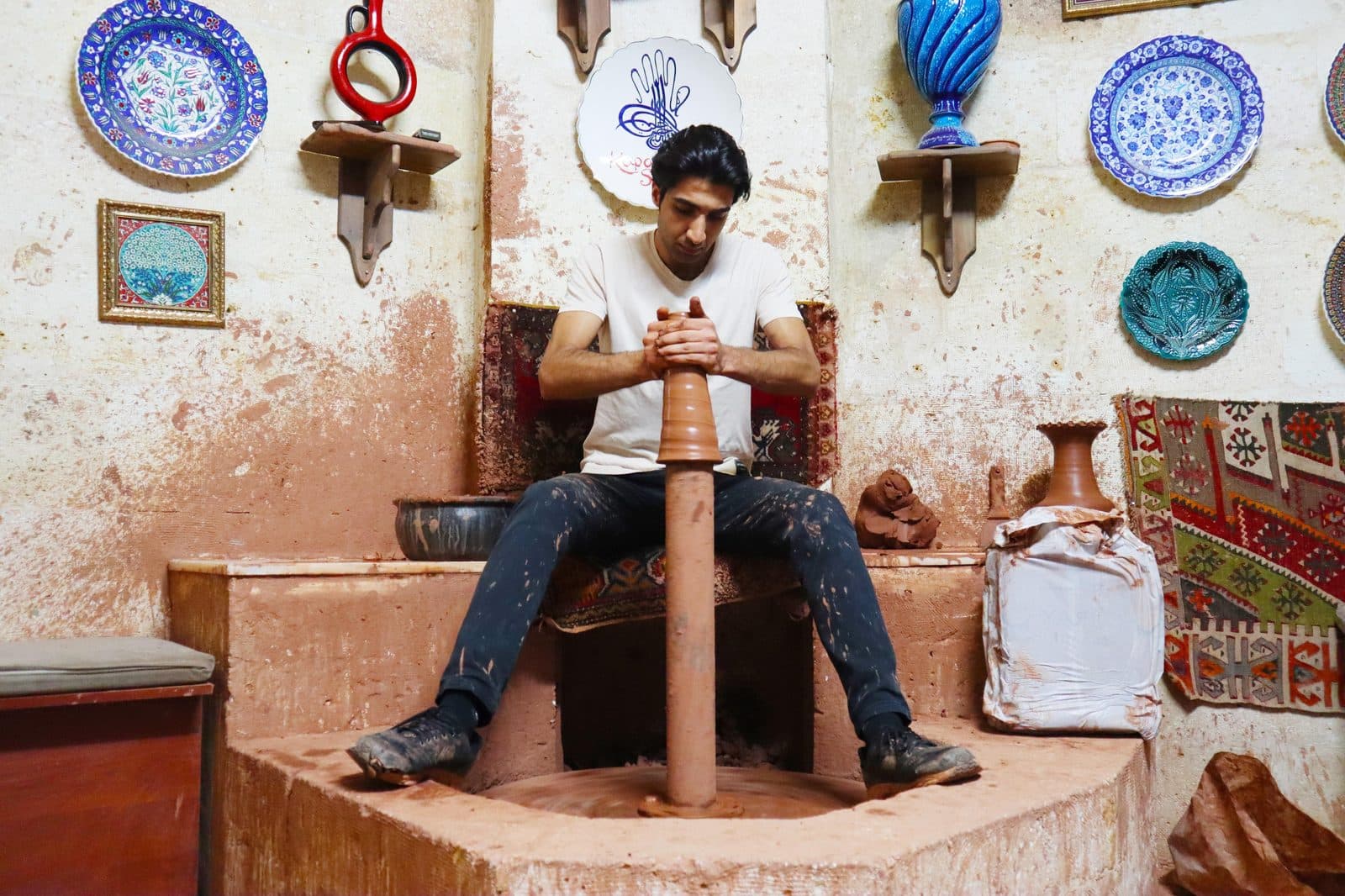 A potter at a wheel, with plates and evil-eye ceramics on the wall. Avanos Pottery Workshop, arranged from The Owl Cave Hotel in Göreme, Cappadocia.
