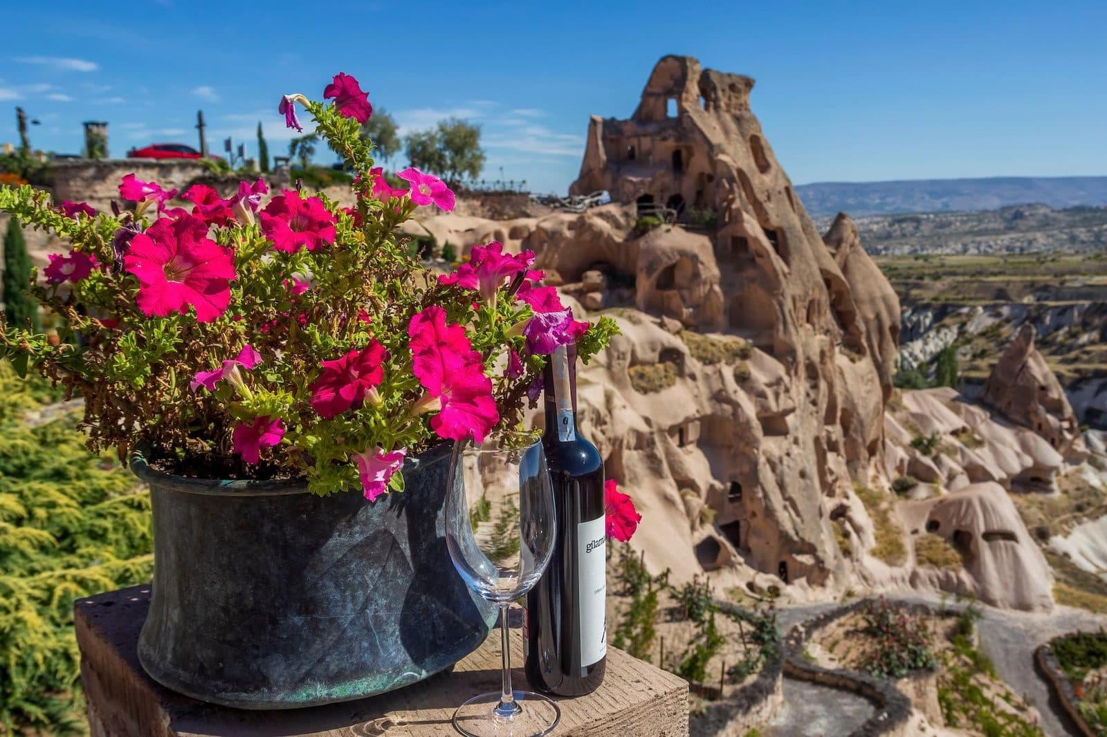 A wine bottle and pink flowers on a ledge, fairy chimneys behind. Avanos Wine Tasting, arranged from The Owl Cave Hotel in Göreme, Cappadocia.