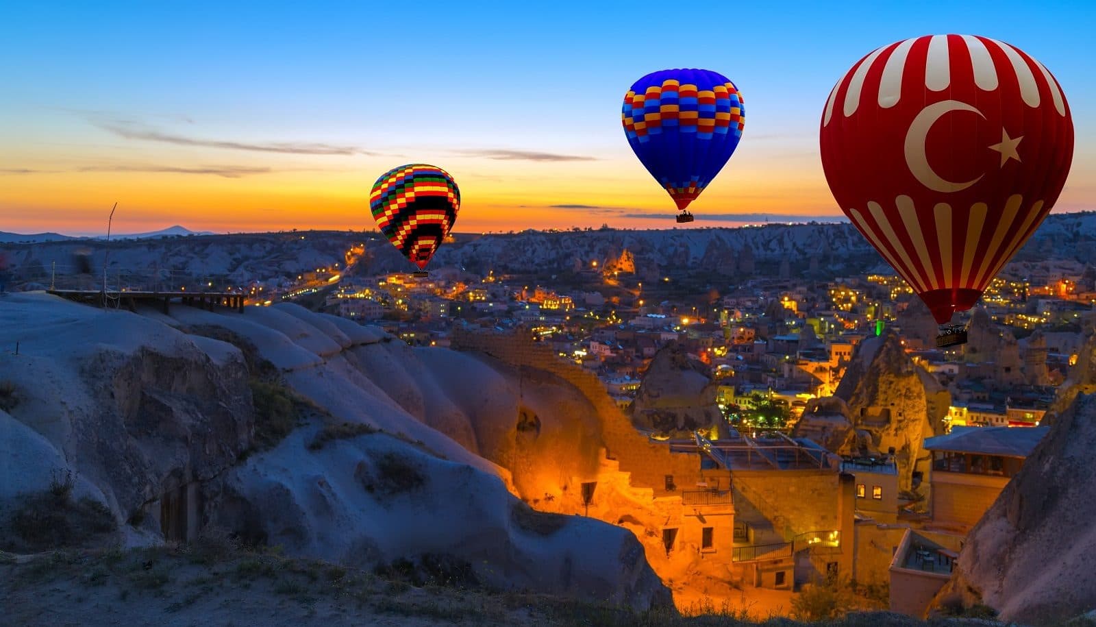Three hot air balloons over a lit town at dusk. Sunrise Balloon and Cappadocia Day, arranged from The Owl Cave Hotel in Göreme, Cappadocia.
