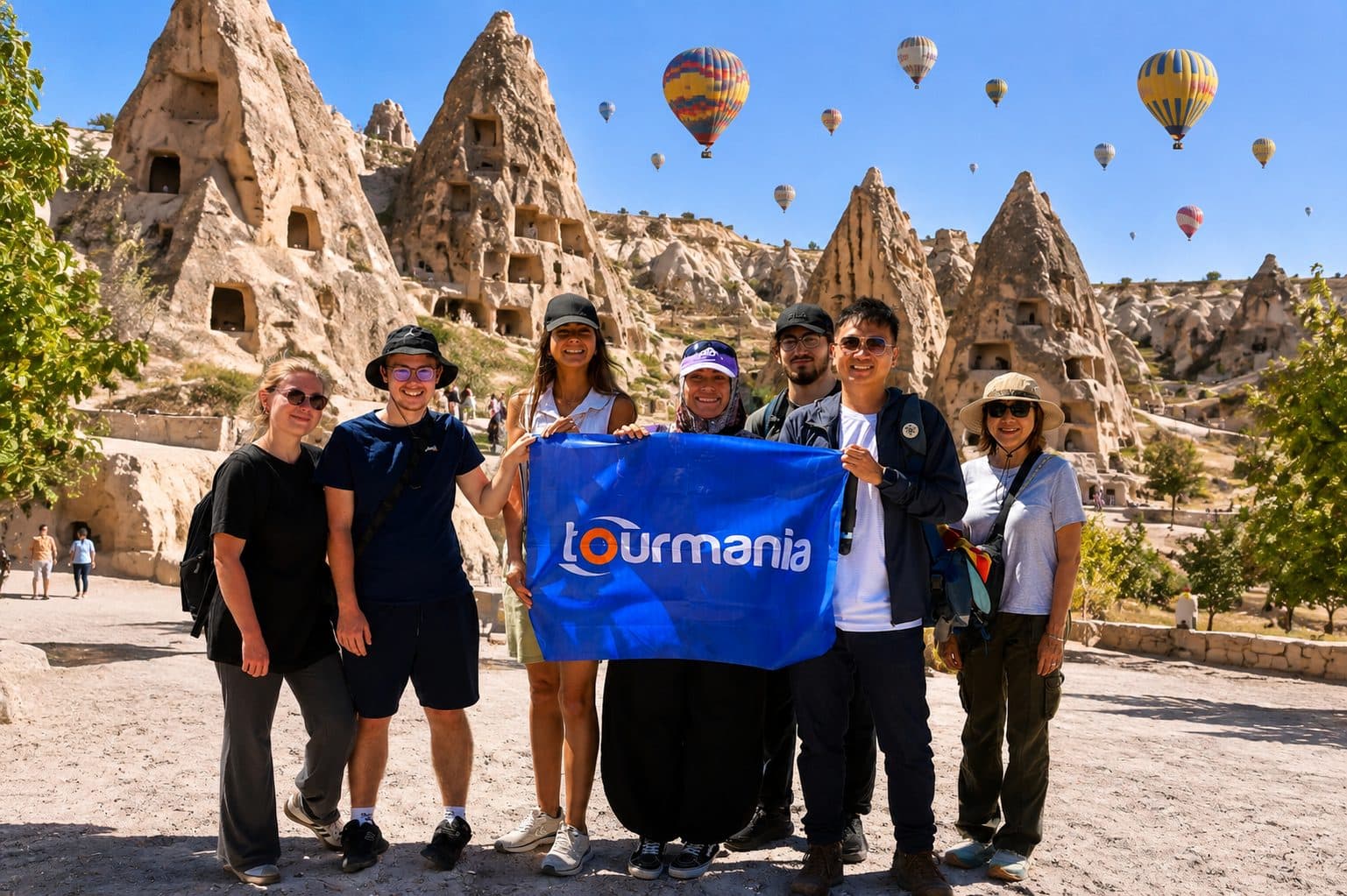 A group holding a Tourmania flag among fairy chimneys and balloons. Balloon Flight with a Choice of Day Tour, arranged from The Owl Cave Hotel in Göreme, Cappadocia.