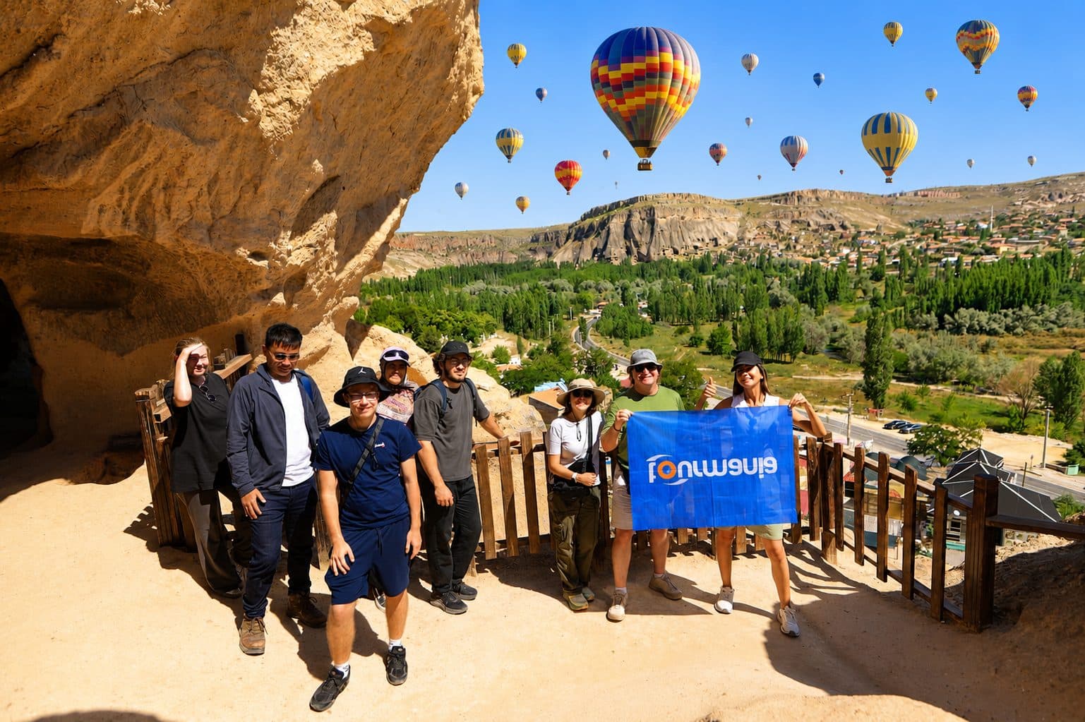 A group with a Tourmania flag at a viewpoint, balloons filling the sky. Balloon Flight with a Choice of Day Tour, arranged from The Owl Cave Hotel in Göreme, Cappadocia.