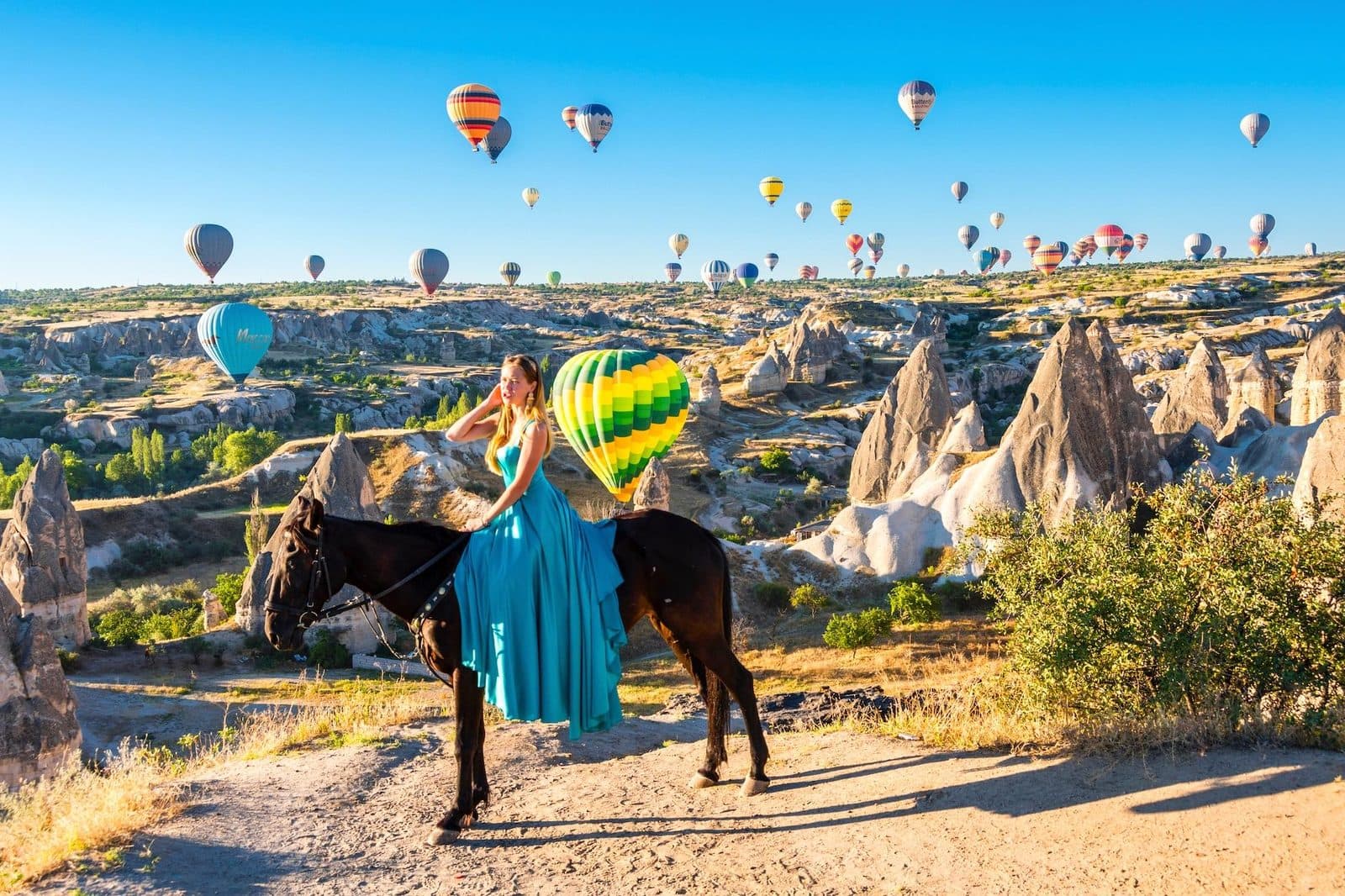 A guest in a blue dress with a dark horse, balloons over the chimneys. Cappadocia from Antalya, arranged from The Owl Cave Hotel in Göreme, Cappadocia.
