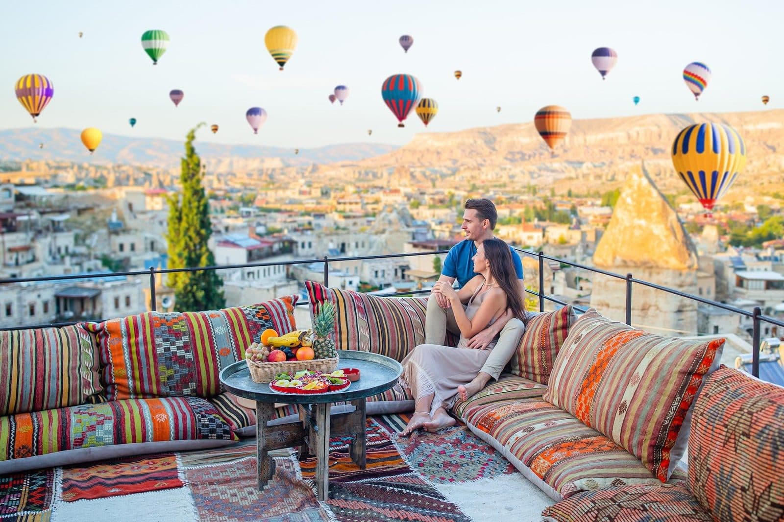 A couple on a terrace overlooking a town and hot air balloons. Cappadocia from Antalya, arranged from The Owl Cave Hotel in Göreme, Cappadocia.