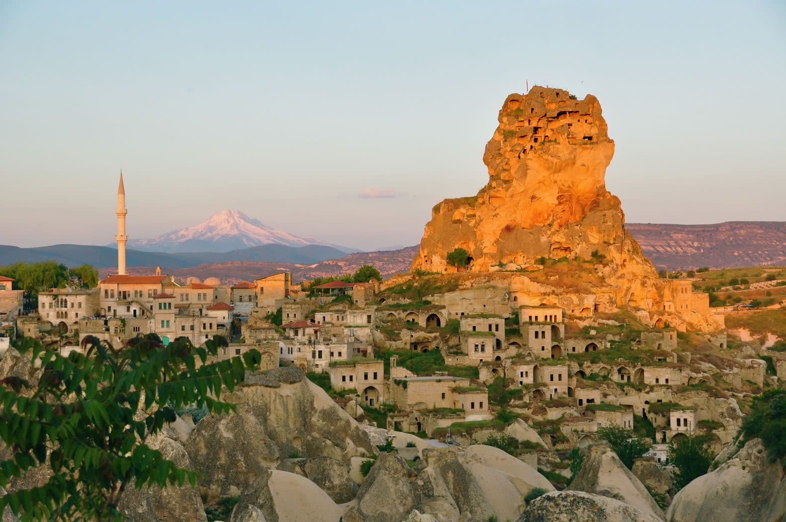 A town and minaret below a rock outcrop, with a snow-capped mountain beyond. Green Tour with Narlıgöl, arranged from The Owl Cave Hotel in Göreme, Cappadocia.