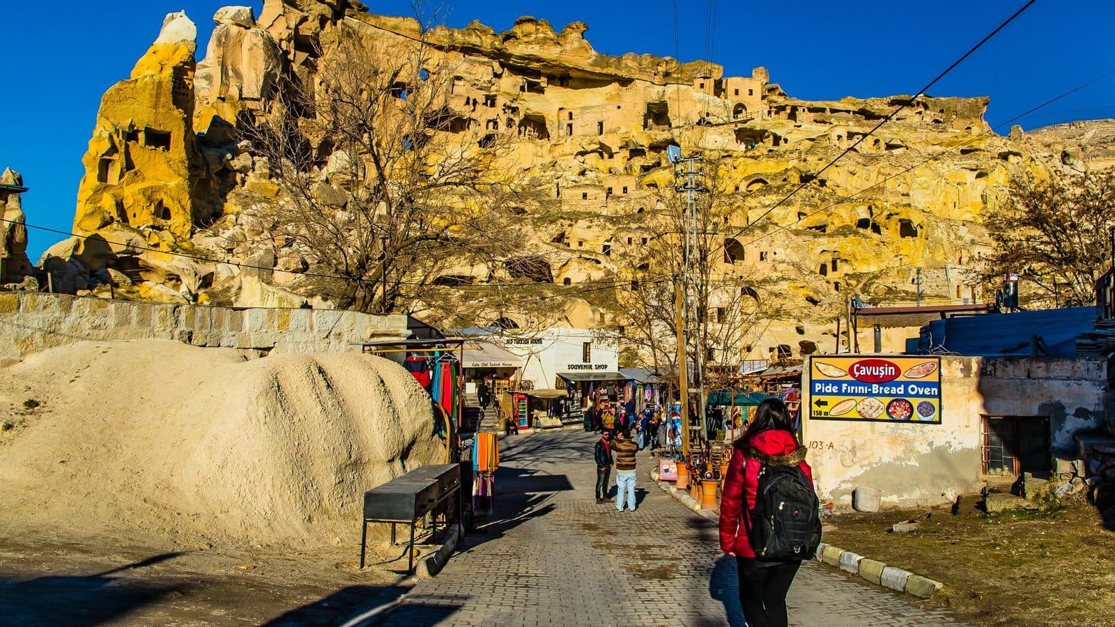 A path toward a cliff of cave rooms, with visitors and small stalls. Cappadocia Red and Green Day, arranged from The Owl Cave Hotel in Göreme, Cappadocia.