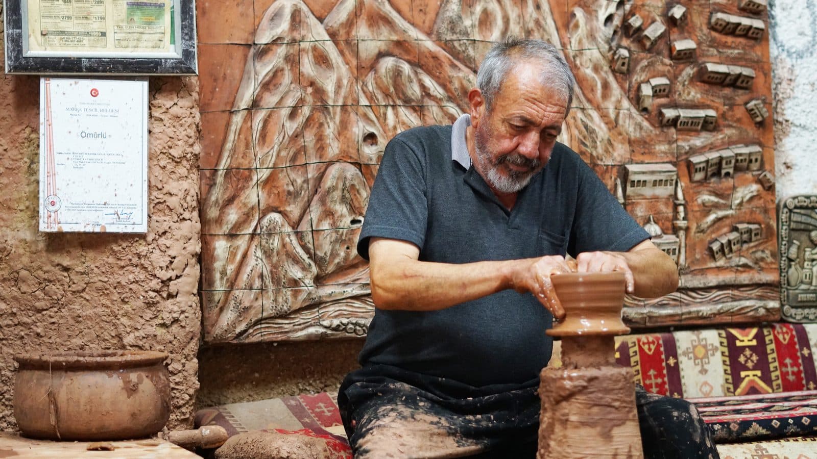 A potter working a kick wheel beside a carved stone wall. Cappadocia Red and Green Day, arranged from The Owl Cave Hotel in Göreme, Cappadocia.
