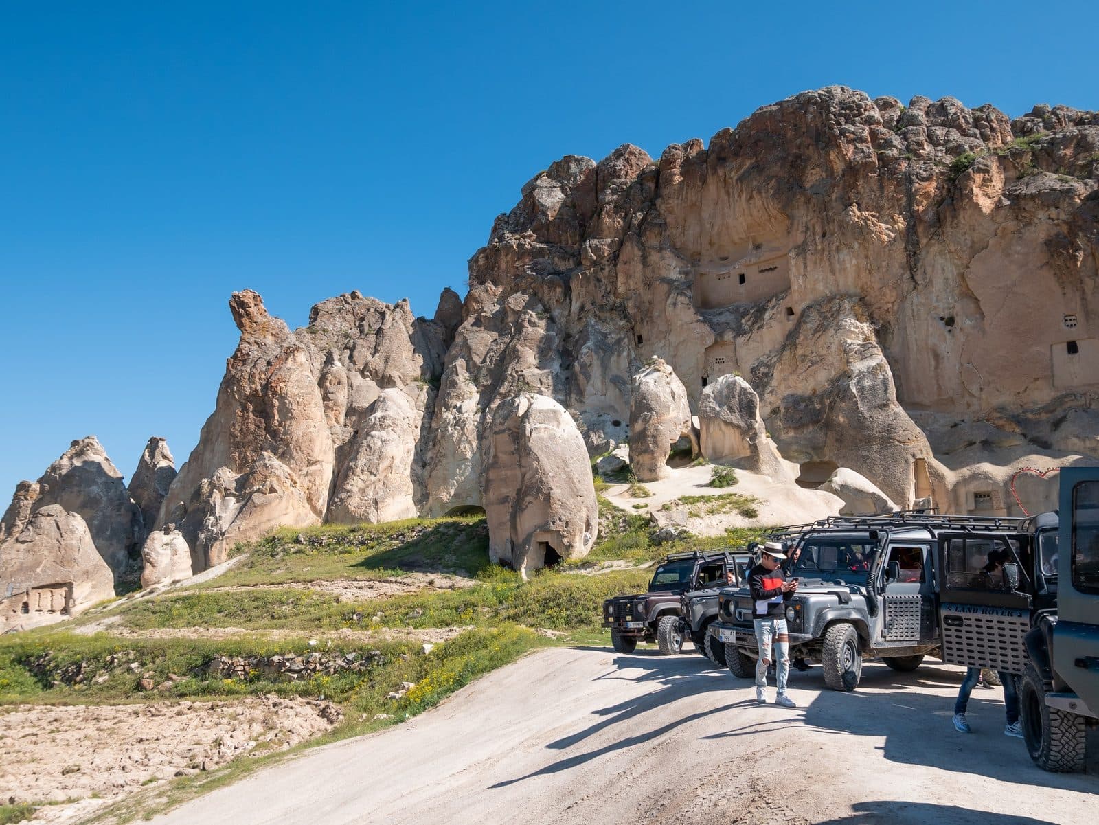 A line of jeeps on a road below a cliff of fairy chimneys. Sunrise or Sunset Jeep Safari, arranged from The Owl Cave Hotel in Göreme, Cappadocia.