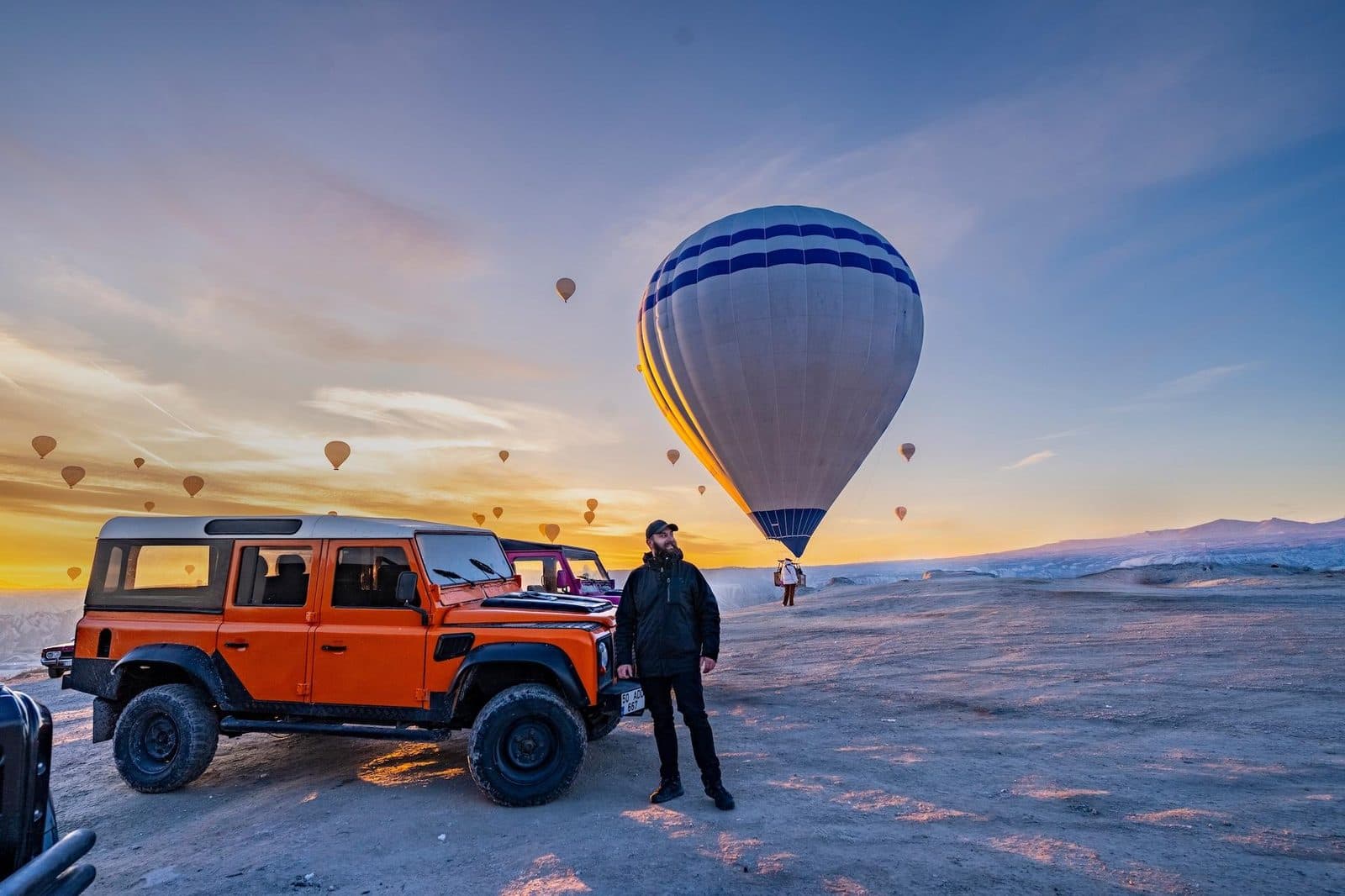 Orange and black jeeps on a ridge at sunrise, with hot air balloons. Sunrise or Sunset Jeep Safari, arranged from The Owl Cave Hotel in Göreme, Cappadocia.