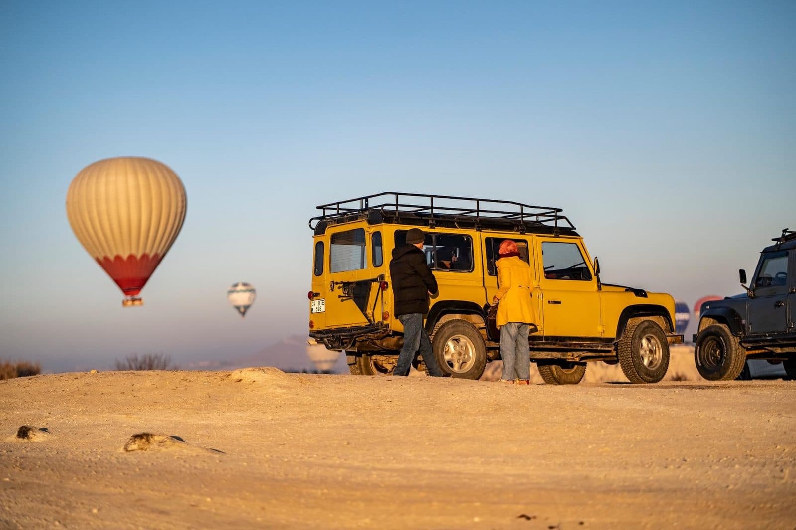 A yellow jeep parked beneath a cream-colored balloon. Sunrise or Sunset Jeep Safari, arranged from The Owl Cave Hotel in Göreme, Cappadocia.