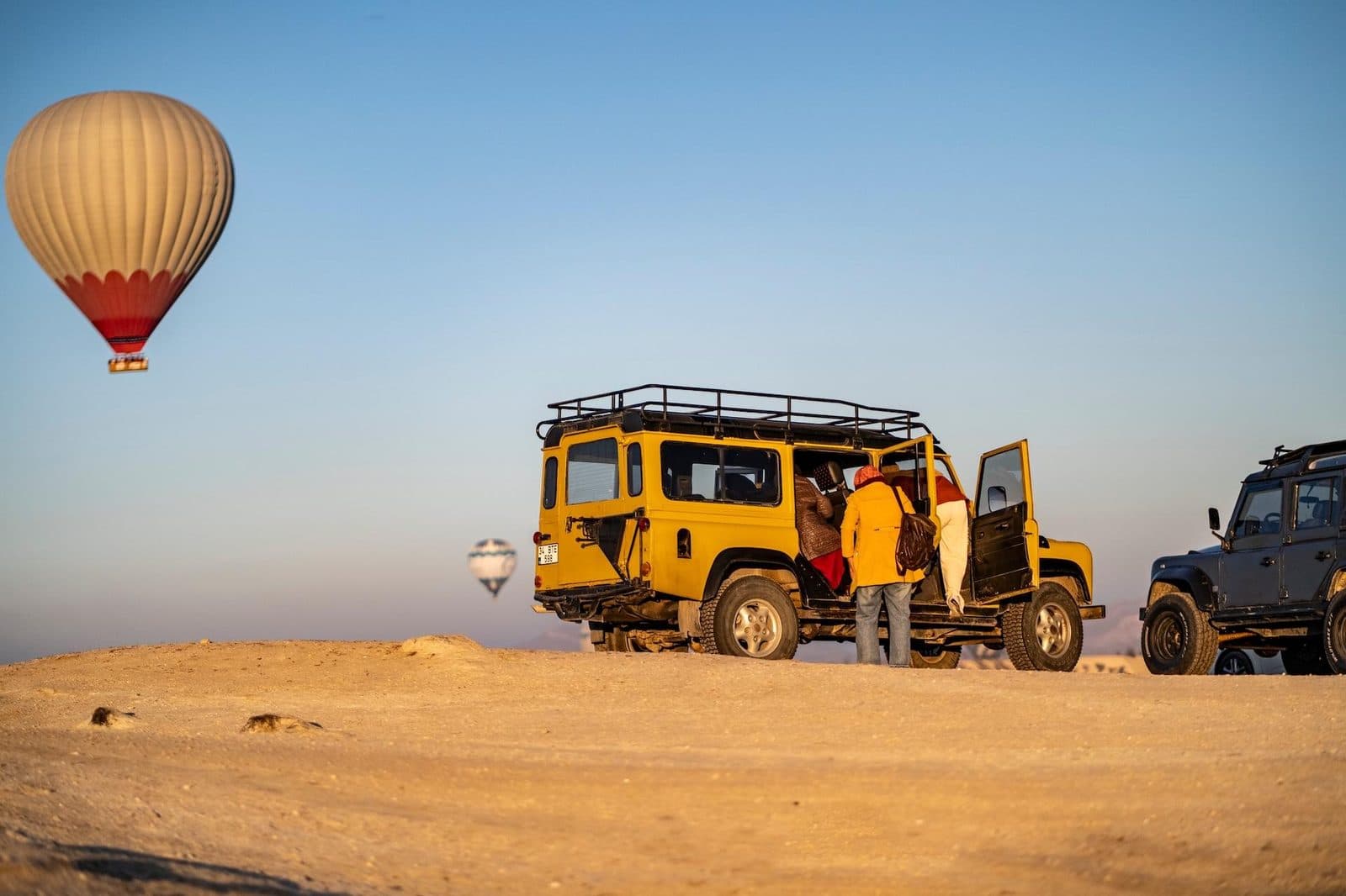 A yellow jeep and its driver on the plateau, a balloon overhead. Sunrise or Sunset Jeep Safari, arranged from The Owl Cave Hotel in Göreme, Cappadocia.