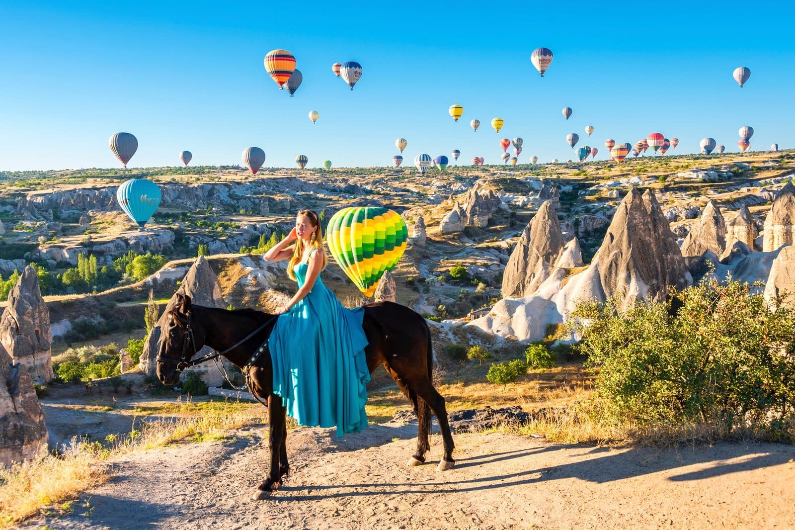 A guest in a blue dress standing with a dark horse among balloons. Sunrise Horse Ride, arranged from The Owl Cave Hotel in Göreme, Cappadocia.
