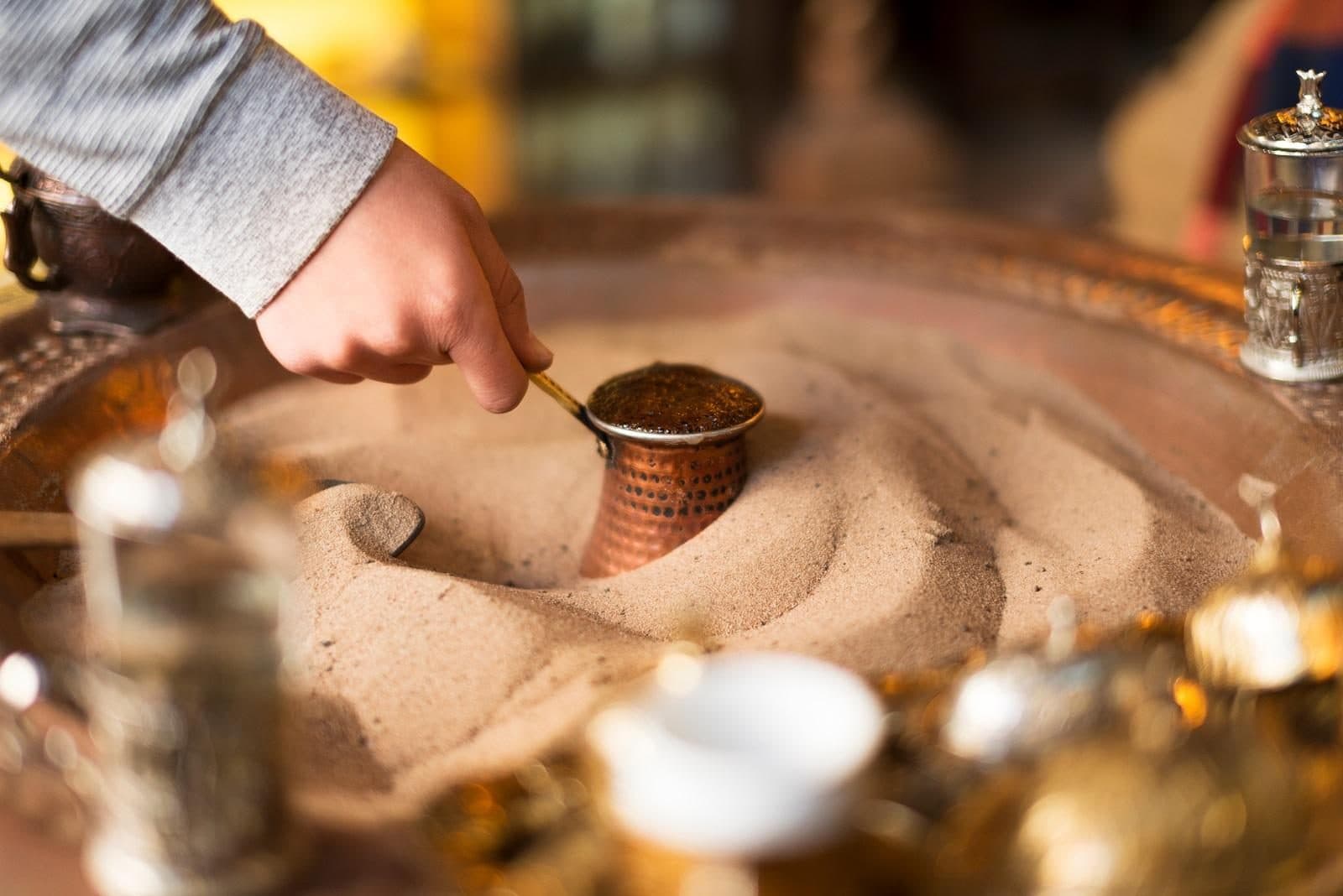 A hand stirring a copper cezve buried in hot sand. Sand-Brewed Turkish Coffee Workshop, arranged from The Owl Cave Hotel in Göreme, Cappadocia.