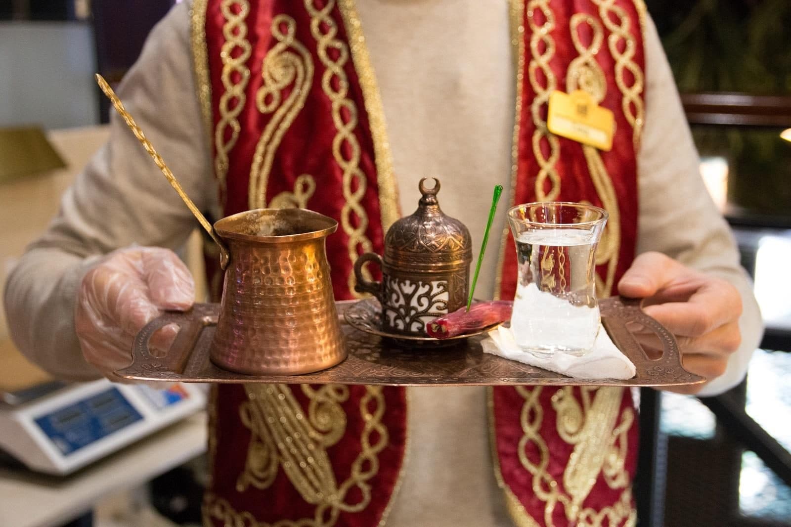 A server in an embroidered vest carrying a tray with a copper cezve. Sand-Brewed Turkish Coffee Workshop, arranged from The Owl Cave Hotel in Göreme, Cappadocia.