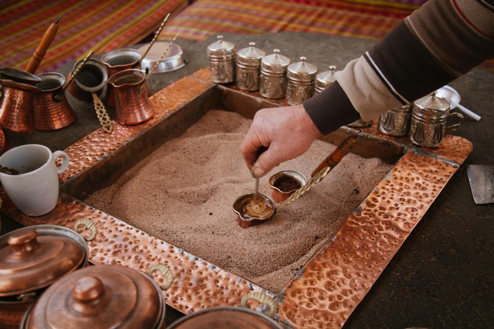 A hand lifting a copper cezve from a sand-filled tray. Sand-Brewed Turkish Coffee Workshop, arranged from The Owl Cave Hotel in Göreme, Cappadocia.