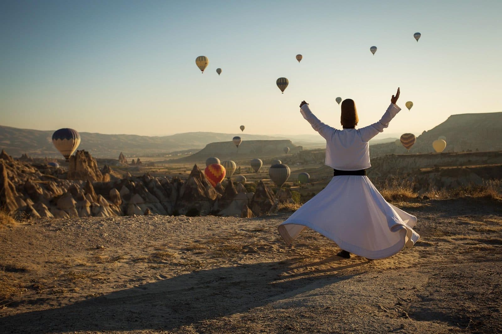 A whirling dervish spinning outdoors at sunrise, balloons in the sky. Whirling Dervish Ceremony and Dinner, arranged from The Owl Cave Hotel in Göreme, Cappadocia.